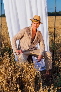 Kırsal kesimde. Kalın keten takım elbiseli, şapkalı uzun boylu yakışıklı bir adam altın yulaf tarlasında eski bir bavulda oturuyor ve arka planda beyaz bir perde var..