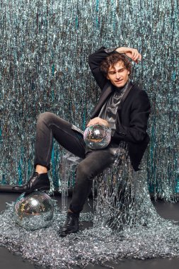 Uzun boylu çekici bir adam, ışıltılı arka planda yerde oturuyor ve ayna disko topu elinde şampanya kadehi tutuyor. Parti konsepti.