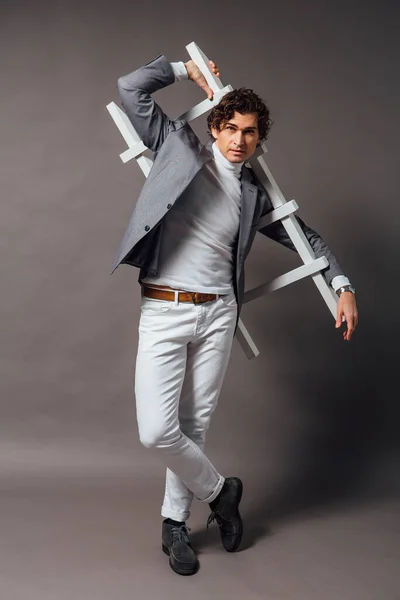 一位高大英俊男子的时尚肖像 身穿白色高领毛衣 白色牛仔裤和灰色夹克 背景灰暗 脚上有白色梯子 — 图库照片