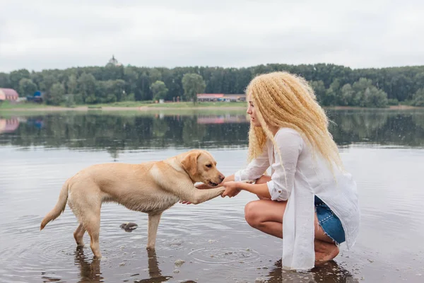 若いです美しいです女性とともにブロンド巻き毛の再生とともに彼女のラブラドール取得犬で川 — ストック写真