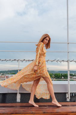 Uzun leopar desenli elbiseli güzel bir kadın. Çıplak ayakla bir binanın çatısında duruyor..
