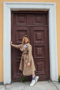 Genç, gülümseyen, vahşi saçlı, sonbahar montu giymiş, eski bir binanın kapısında poz veren bir kadın..