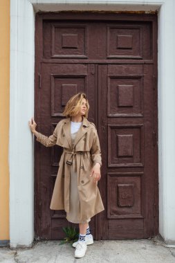 Genç, gülümseyen, vahşi saçlı, sonbahar montu giymiş, eski bir binanın kapısında poz veren bir kadın..