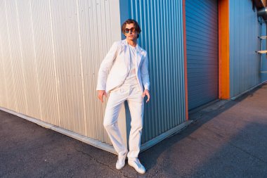 Gün batımında binanın köşesinde duran beyaz takım elbiseli ve yuvarlak güneş gözlüklü yakışıklı bir adamın geniş açılı portresi.