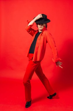 Kırmızı gömlekli, siyah şapkalı, kırmızı arka planda poz veren uzun boylu yakışıklı bir adamın portresi..