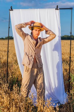 Kırsal kesimde. Uzun boylu, keten takım elbiseli ve şapkalı yakışıklı bir adam altın yulaf tarlasında dikiliyordu. Arka planda beyaz bir perde vardı..