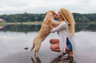 Sarı kıvırcık saçlı genç bir kadın nehir kıyısında Labrador av köpeğiyle sarmaş dolaş.