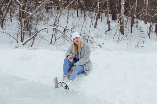 アイススケートに乗ってリラックスした若い女性や アイスリンクのポットで熱々のドリンクを飲む 女の子は雪の上に座って笑顔 — ストック写真