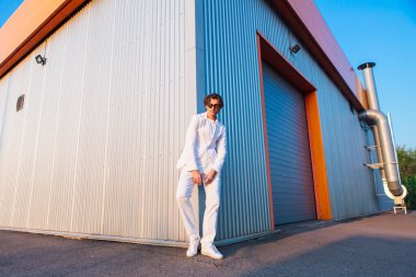 Gün batımında binanın köşesinde duran beyaz takım elbiseli ve yuvarlak güneş gözlüklü yakışıklı bir adamın geniş açılı portresi.