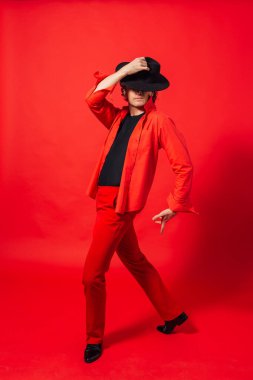 Kırmızı gömlekli, siyah şapkalı, kırmızı arka planda poz veren uzun boylu yakışıklı bir adamın portresi..