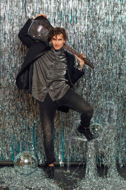 Parlak arka planda elektro gitar tutan uzun boylu çekici bir adam ve yerde ayna disko topu var. Parti konsepti.