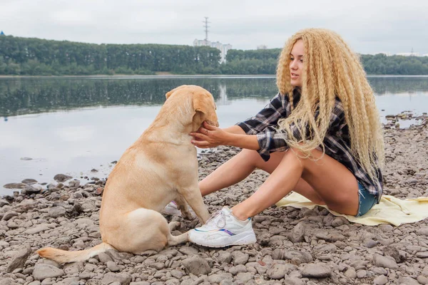 若いです美しいです女性とともにブロンド巻き毛で座っています彼女のラブラドール取得犬オンザ川の海岸 — ストック写真