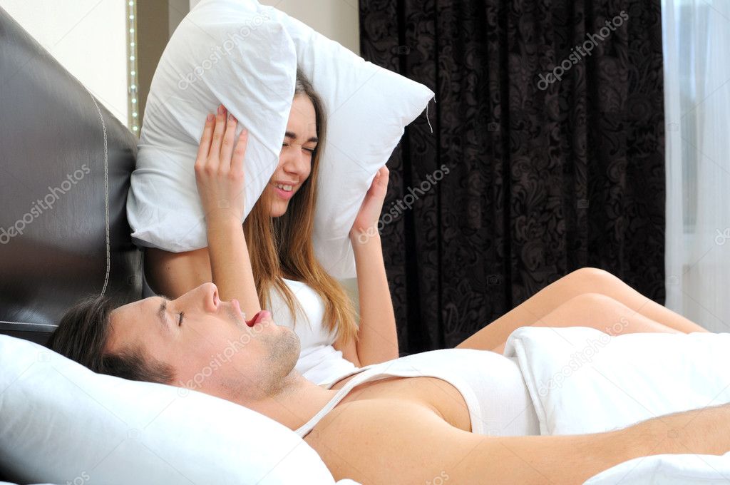 Scenic in bedroom of loving young couple lying in bed and girl holding pillow on the ears because of snoring boyfriend