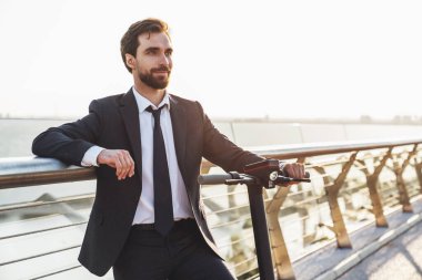 Şehrin arka planında elektrikli scooterla gezerken iş elbisesi giymiş sakallı genç bir işadamı.