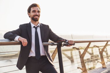 Şehrin arka planında elektrikli scooterla gezerken iş elbisesi giymiş sakallı genç bir işadamı.