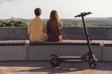 Genç bir çift sırtı kameraya dönük bir bankta oturuyor ve şehrin manzarasına hayranlıkla bakarken şehrin arka planında elektrikli scooterla geziyor..