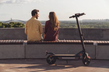 Genç bir çift sırtı kameraya dönük bir bankta oturuyor ve şehrin manzarasına hayranlıkla bakarken şehrin arka planında elektrikli scooterla geziyor..