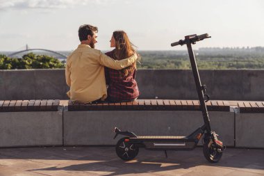 Genç bir çift sırtı kameraya dönük bir bankta oturuyor ve şehrin manzarasına hayranlıkla bakarken şehrin arka planında elektrikli scooterla geziyor..