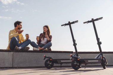 Birkaç genç turist sabah şehirde elektrikli scooter sürerken bankta oturup kahve içerek sohbet ediyorlar..