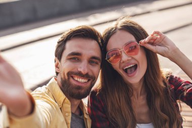 Genç çift bankta konuşarak ve selfie çekerek eğleniyor. Sabah şehirde yürürken telefonla..