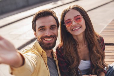 Genç çift bankta konuşarak ve selfie çekerek eğleniyor. Sabah şehirde yürürken telefonla..