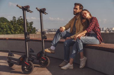 Genç bir çift bankta oturuyor ve rahatlıyor. Sabahları elektrikli scooterlara biniyorlar..