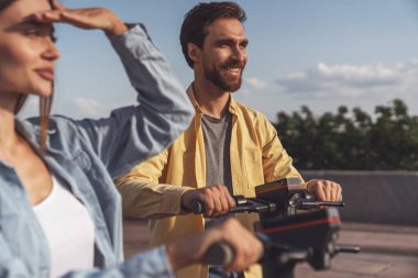 Genç ve neşeli bir çift yazın güneşli bir havada şehir parkında elektrikli scooter kullanıyor..