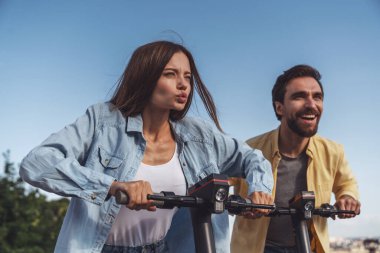 Genç ve neşeli bir çift yazın güneşli bir havada şehir parkında elektrikli scooter kullanıyor..
