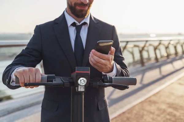電気スクーターに乗っている間 ビジネススーツの若い陽気な髭のビジネスマンは 朝の街の背景にスマートフォンを使用しています — ストック写真