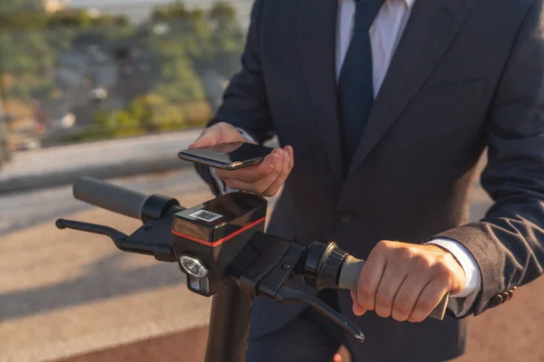 電気スクーターに乗っている間 ビジネススーツの若い陽気な髭のビジネスマンは 朝の街の背景にスマートフォンを使用しています — ストック写真