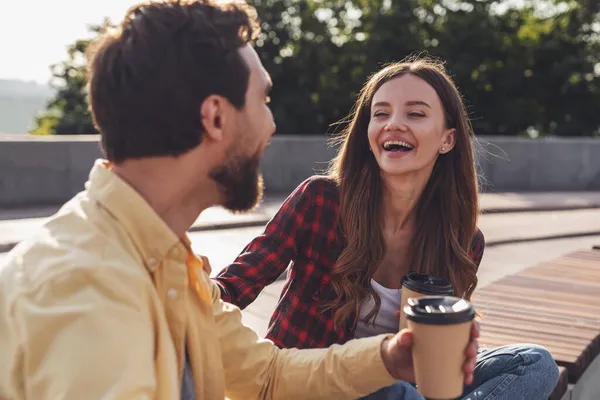 朝の街を歩きながらベンチに座ってコーヒーを飲みながら話す観光客の若いカップル — ストック写真