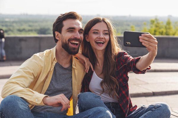 Young couple having fun talking on a bench and taking a selfie using the phone while walking in the morning city.