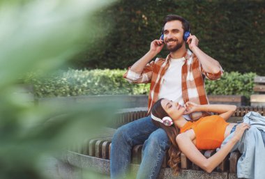 Genç ve neşeli bir çift, parktaki bankta oturmuş kulaklıkla müzik dinliyor. Kız adamın ayaklarının dibinde yatıyor..