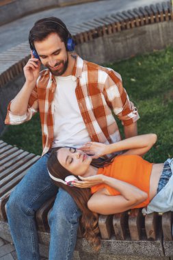 Genç ve neşeli bir çift, parktaki bankta oturmuş kulaklıkla müzik dinliyor. Kız adamın ayaklarının dibinde yatıyor..