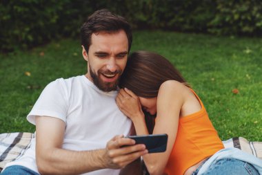 Genç, neşeli bir çift piknik sırasında parkta battaniyeye uzanıyor ve telefonla selfie çekiyor..