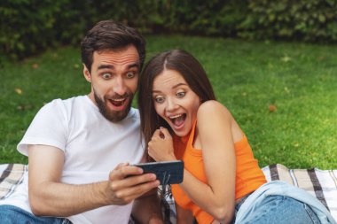 Genç, neşeli bir çift piknik sırasında parkta battaniyeye uzanıyor ve telefonla selfie çekiyor..