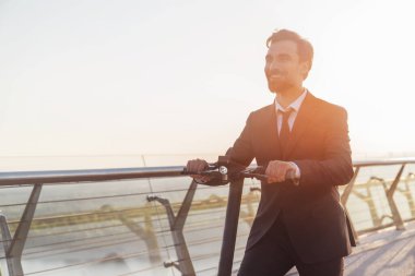 Şehrin arka planında elektrikli scooterla gezerken iş elbisesi giymiş sakallı genç bir işadamı.