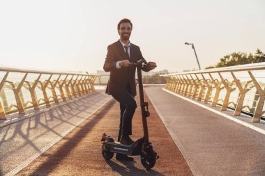 Şehrin arka planında elektrikli scooterla gezerken iş elbisesi giymiş sakallı genç bir işadamı.