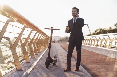 Elektrikli scooter sürerken iş adamı kılığındaki genç, sakallı iş adamı kahve içer ve şehrin arka planında akıllı telefon kullanır.