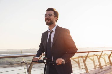 Şehrin arka planında elektrikli scooterla gezerken iş elbisesi giymiş sakallı genç bir işadamı.