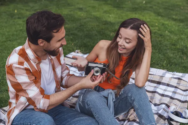 ピクニック中に公園の毛布の上に座っている若い陽気なカップルと古いカメラでお互いの写真を撮る — ストック写真