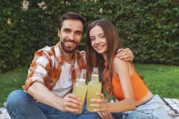 ピクニック中に公園の毛布の上に座って ボトルからアルコールを飲む若い陽気なカップル — ストック写真