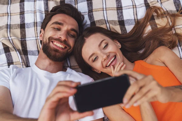 ピクニック中に公園の毛布の上に寝そべっている若い陽気なカップルと携帯電話を使って自撮り — ストック写真