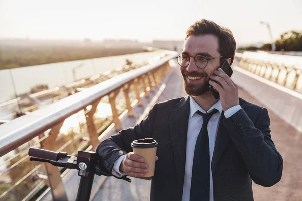 留着胡子 身穿西装的商人坐在电动车上 一边微笑着 一边喝着咖啡 一边在早上的城市里用智能手机 — 图库照片