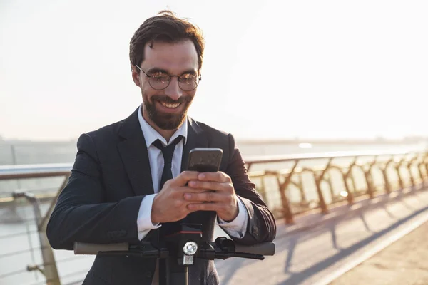 電気スクーターに乗っている間 ビジネススーツの若い陽気な髭のビジネスマンは 朝の街の背景にスマートフォンを使用しています — ストック写真