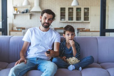 Mutlu küçük bir çocuk ve neşeli babası birlikte televizyon karşısında oturmuş, patlamış mısır yiyor ve evlerindeki en sevdikleri futbol takımı için neşeyle tezahürat yapıyorlar..