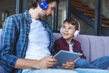 Mutlu küçük çocuk ve neşeli babası evde oturma odasındaki kanepede oturan aletleri kullanarak kulaklıkla müzik dinliyorlar..