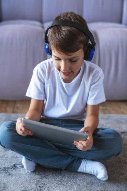 Kulaklıkla müzik dinleyen ve evdeki oturma odasında yerde oturmuş elektronik tablet kullanan küçük bir çocuğun portresi..