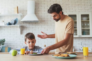 Yemek yemek istemeyen küçük bir çocuğun portresi ve kızgın babası ona evde mutfakta kahvaltı ederken masada yemek yediriyor..