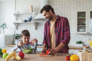 Neşeli baba ve küçük oğlu mutfaktaki masada oturmuş salata hazırlarken birbirlerine yardım ediyorlar..